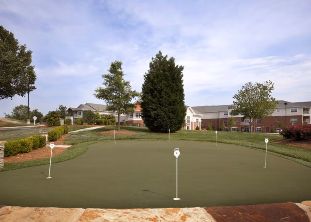 Outdoor putting green with several golf holes and flags, surrounded by grass, trees, and shrubs. In the background, there are multi-story residential buildings under a partly cloudy sky.