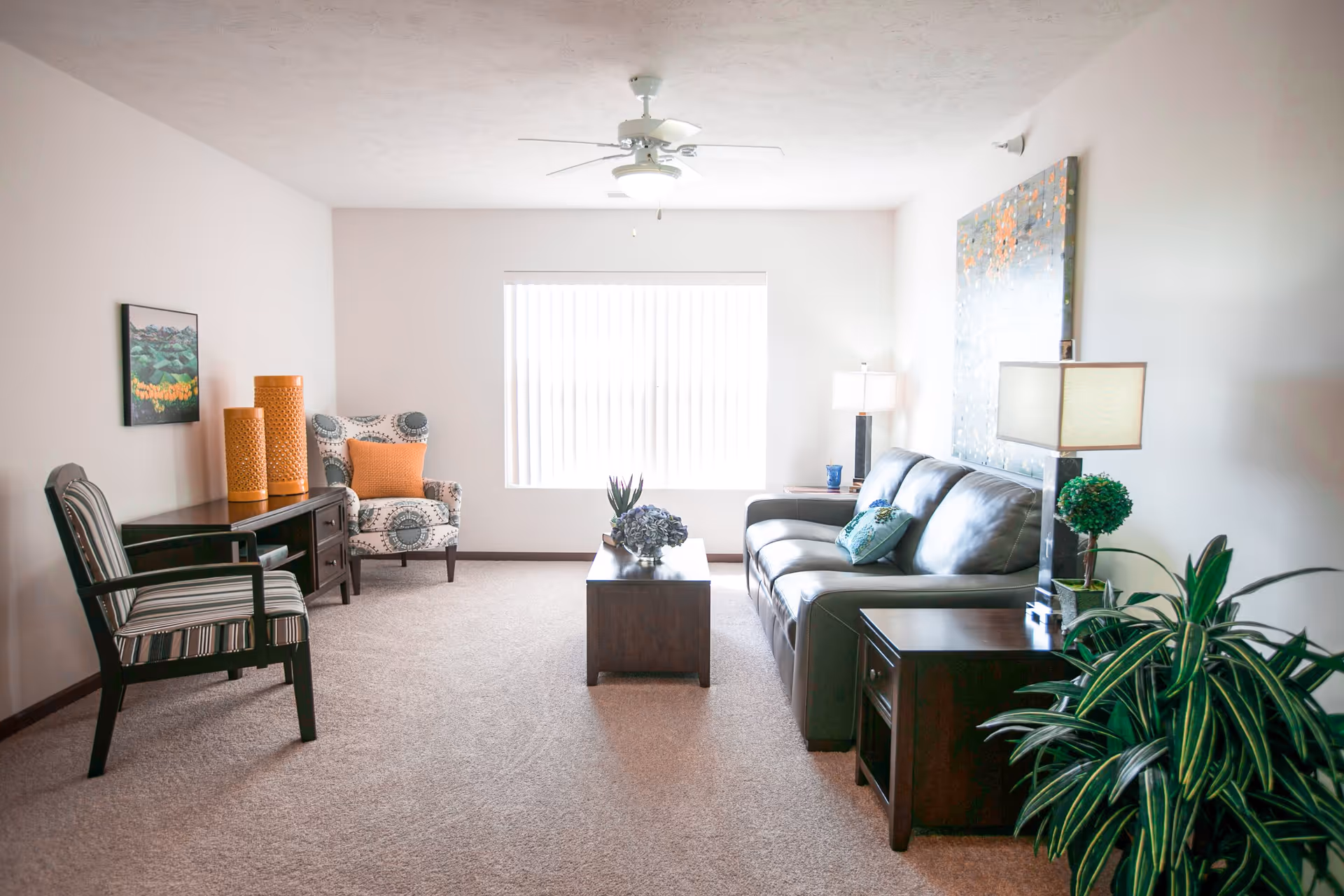 A bright living room with a large window covered by vertical blinds. The room features a dark leather sofa with decorative pillows, a wooden coffee table with a floral centerpiece, two side tables with lamps, a patterned armchair with an orange pillow, a wooden chair with striped upholstery, a wooden console table with decorative orange vases, a framed landscape painting on the wall, a ceiling fan with a light, and a green potted plant in the foreground.