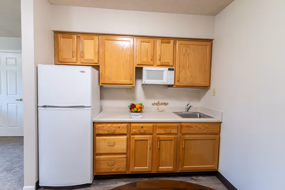 A small kitchen area with wooden cabinets, a white refrigerator, a white microwave mounted above the counter, a stainless steel sink, and a bowl of fruit on the countertop. There is also a decorative sign that says 'welcome home'.