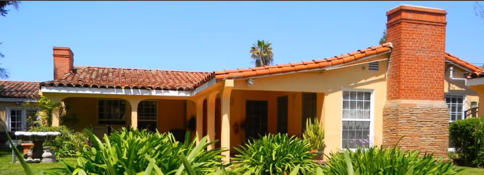 Exterior view of a single-story senior living facility building with a red tile roof, yellow walls, large windows, and a prominent brick chimney. The foreground features lush green plants and a well-maintained lawn under a clear blue sky.