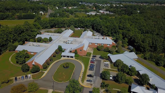 Aerial view of Elizabeth House Assisted Living facility showing multiple connected buildings with gray roofs surrounded by green trees and lawns. There is a circular driveway with parked cars and a parking lot adjacent to the buildings.