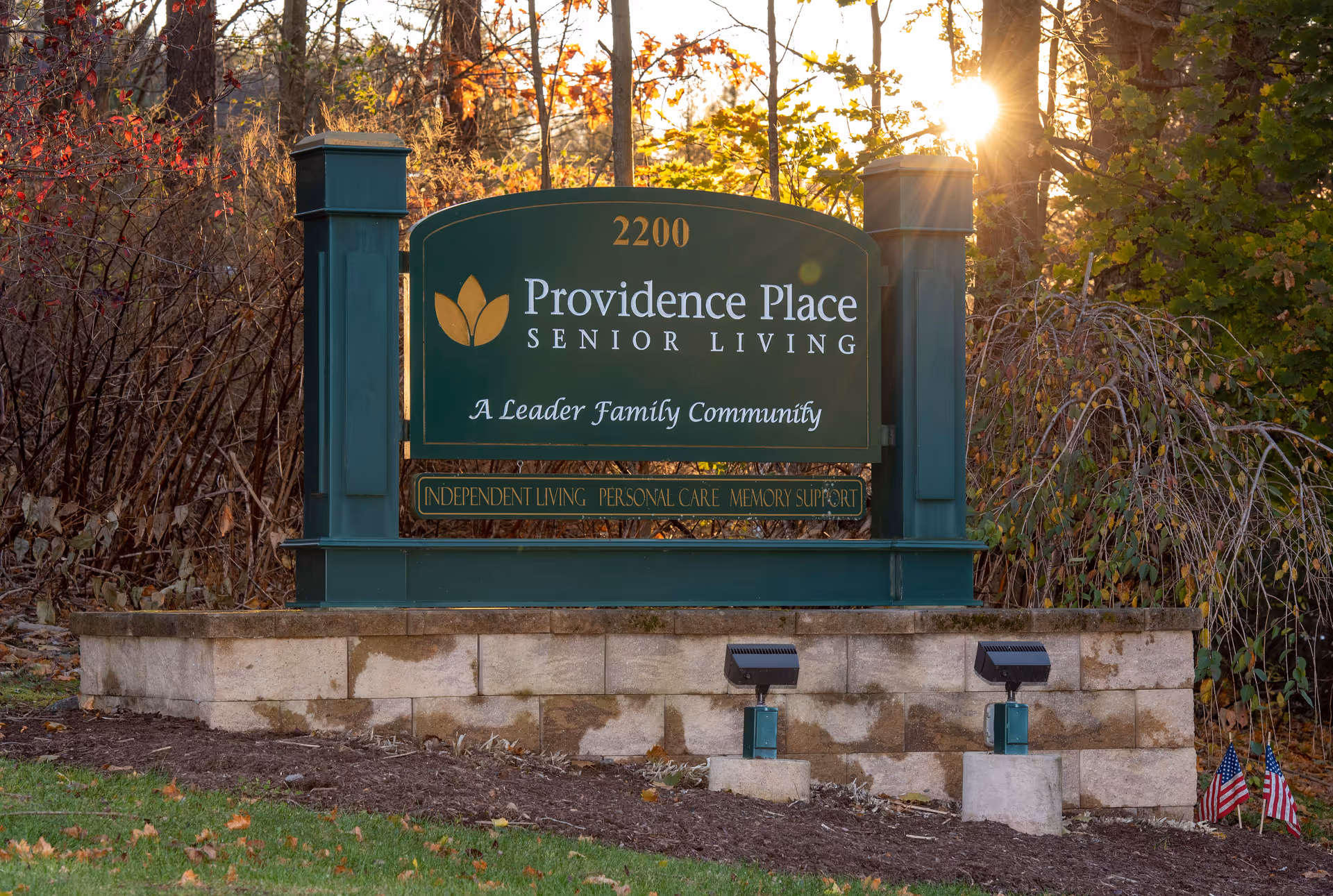 Green entrance sign reading "Providence Place Senior Living" set on a low stone wall with landscaping and sunlight behind it.