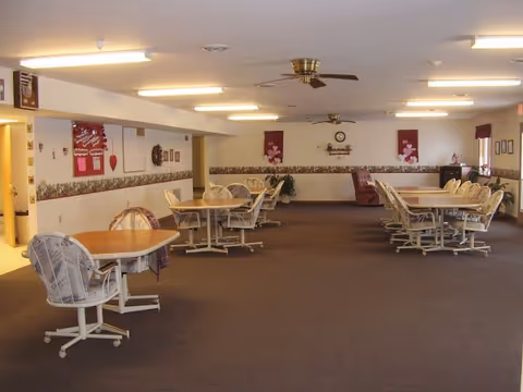 A spacious dining room with several wooden tables and cushioned chairs on wheels arranged neatly. The room has beige walls with a floral border, ceiling fans, fluorescent lighting, and decorative wall hangings including a clock and framed pictures. There is a recliner chair in the back corner and a small plant near the wall.