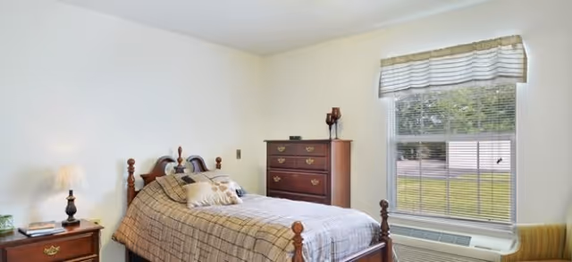 A bedroom with a wooden bed frame and a neatly made bed with patterned bedding and pillows. There is a wooden nightstand with a lamp and a small plant on it to the left of the bed. A wooden dresser with decorative items on top is against the wall. A window with blinds and a valance lets in natural light, and an air conditioning unit is installed below the window. The walls are painted light beige.