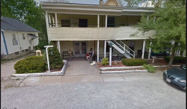 Exterior view of a two-story residential building with a covered porch and stairs leading to the second floor. There are bushes and small trees in front of the building, a paved driveway, and a parked car on the right side. Two people are sitting on the porch.