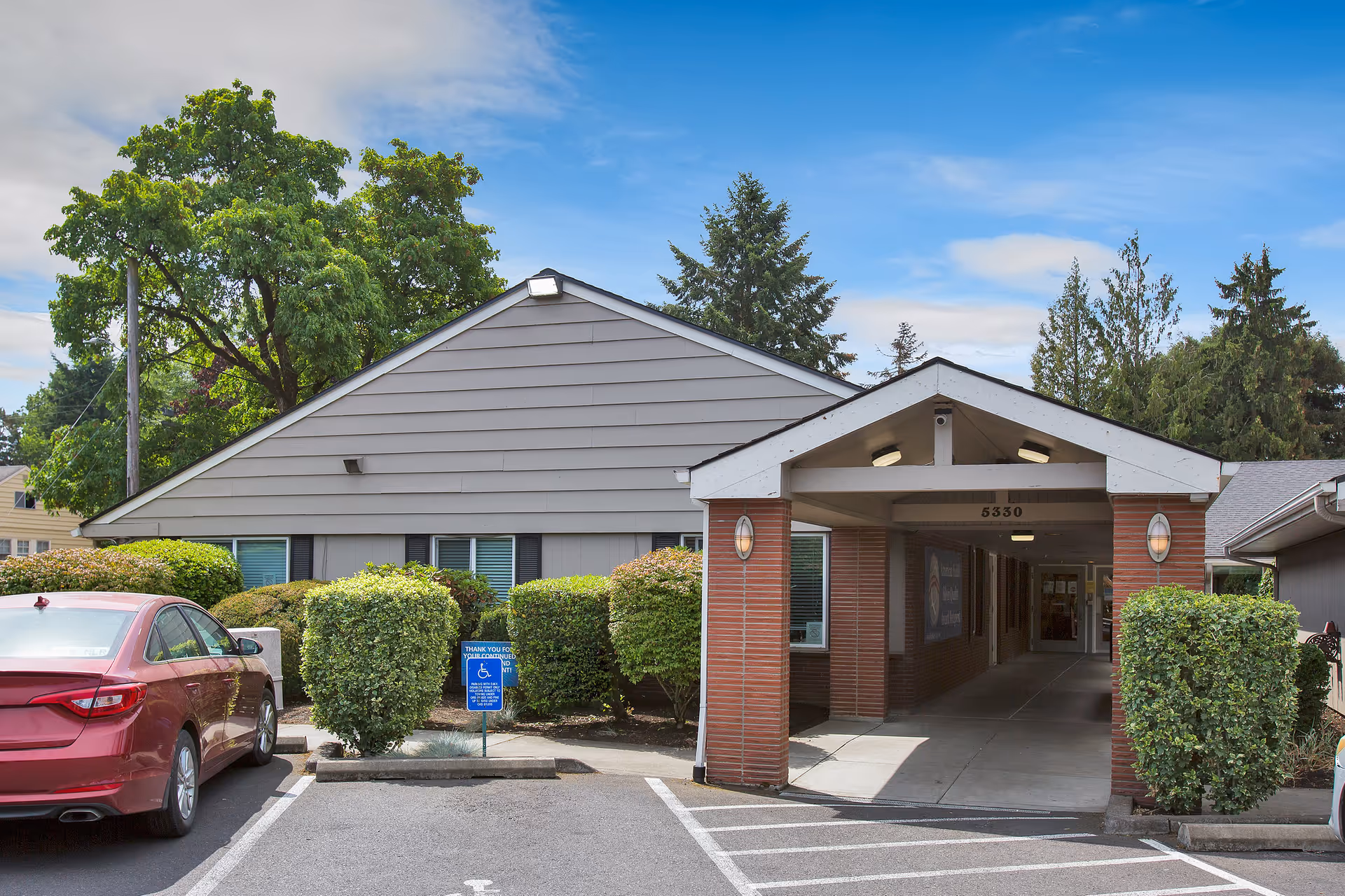 Front entrance of a single-story senior living facility with a covered driveway, shrubs and parked cars.