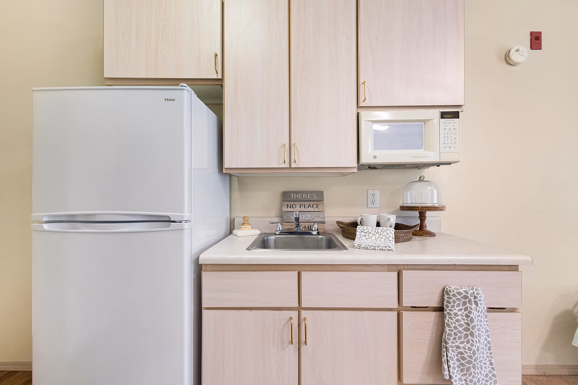 Small kitchen area with light wood cabinets, a white refrigerator, a stainless steel sink, a microwave mounted above the counter, and various kitchen items including a towel hanging from a drawer, a cake stand with a glass cover, two mugs, and a decorative sign that reads 'THERE'S NO PLACE LIKE HOME'.