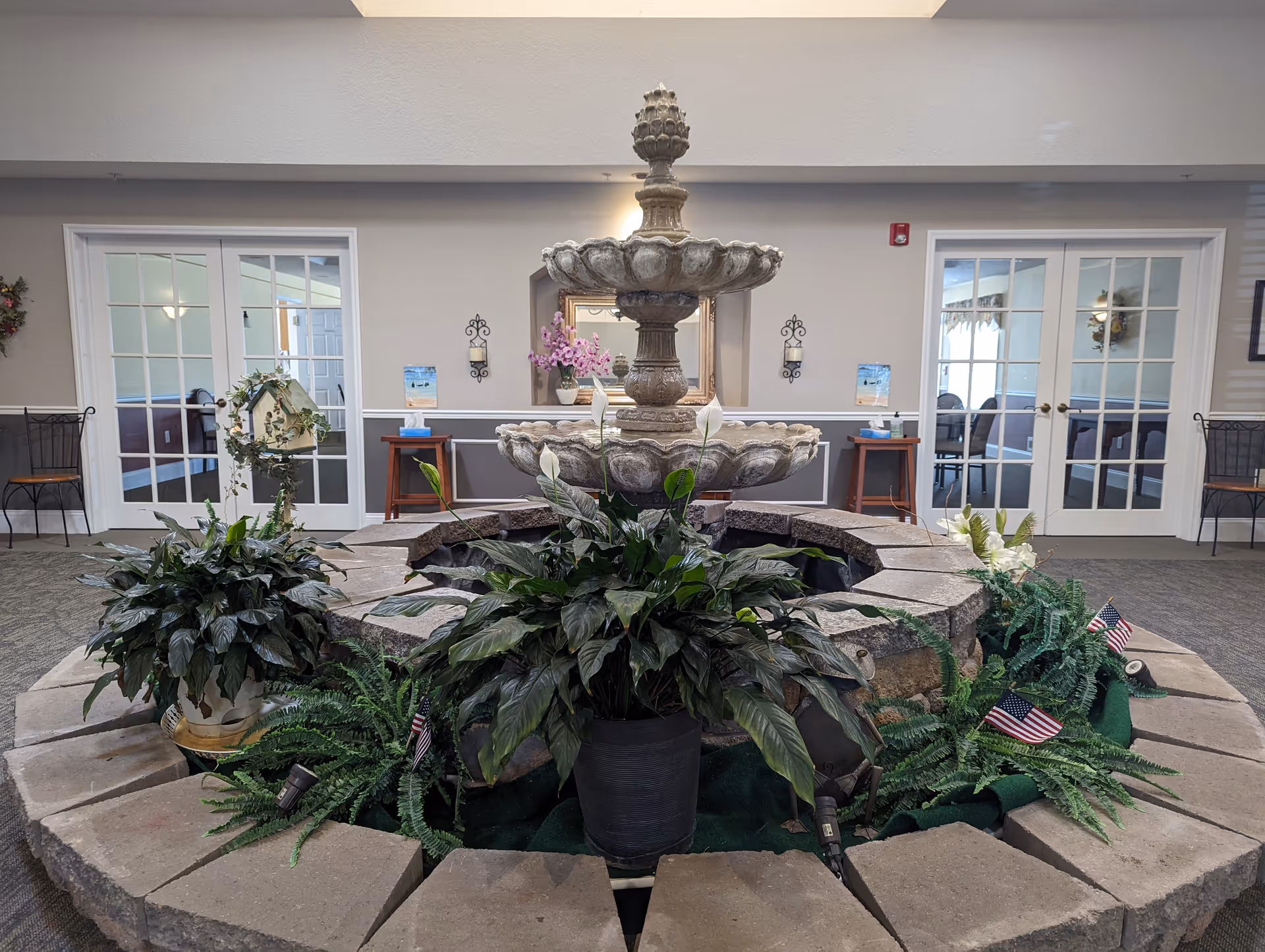 Indoor area with a large stone fountain surrounded by green plants and small American flags. Behind the fountain are two sets of white French doors leading to other rooms, with chairs and tables visible through the glass. The walls are decorated with a mirror, wall sconces, and a small table with flowers.