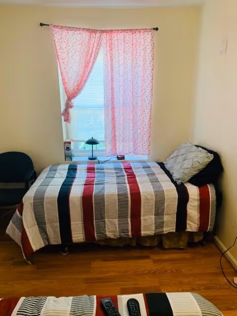 A small bedroom with a single bed covered in a striped bedspread in shades of red, black, gray, and white. There is a window with pink patterned curtains partially drawn, a small black lamp on the windowsill, and a black chair next to the bed. The floor is wooden.