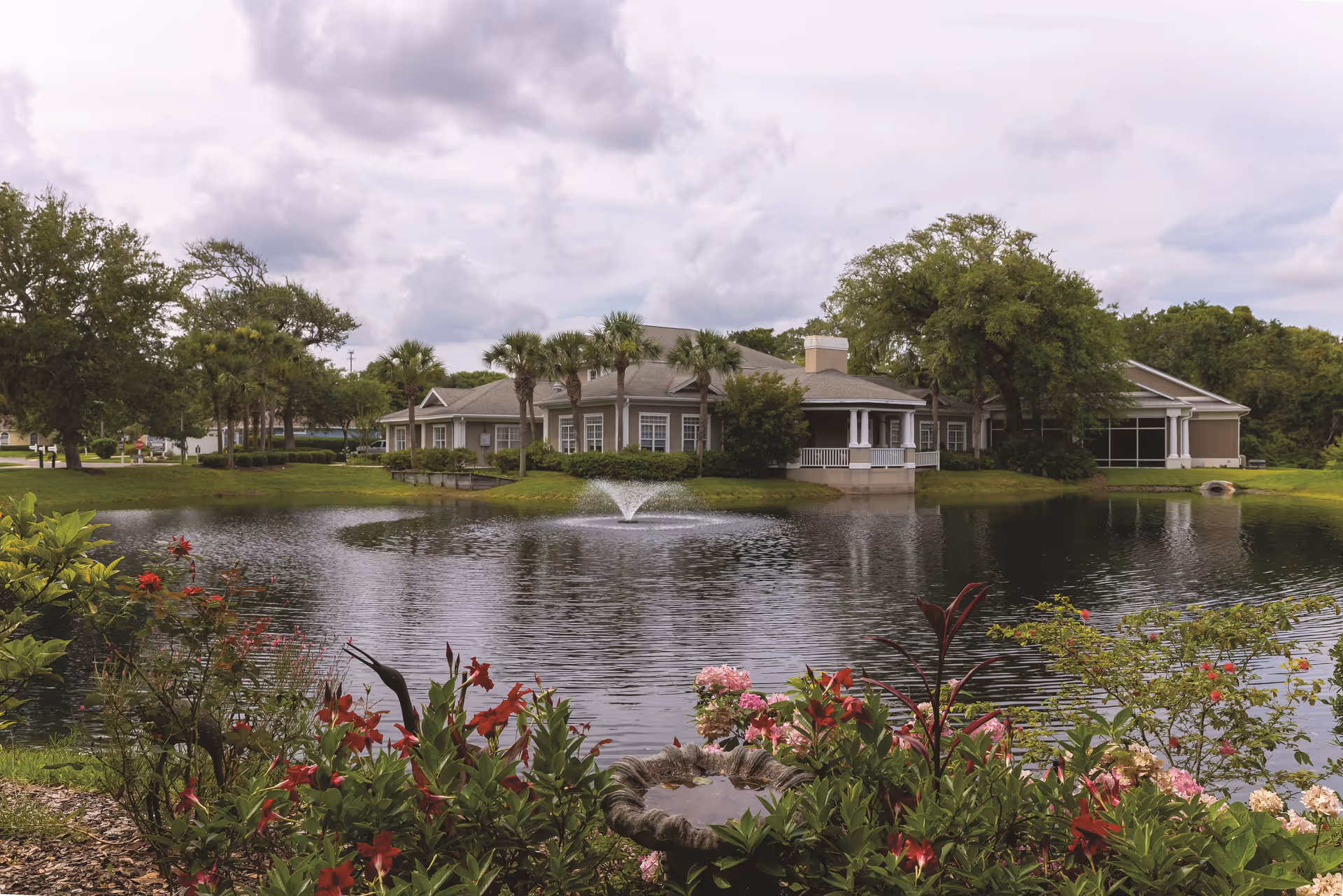 View of a senior living facility building named Retreat At Osprey Village, seen across a pond with a water fountain in the center. The foreground features colorful flowers and greenery, with palm trees and other trees surrounding the building under a cloudy sky.