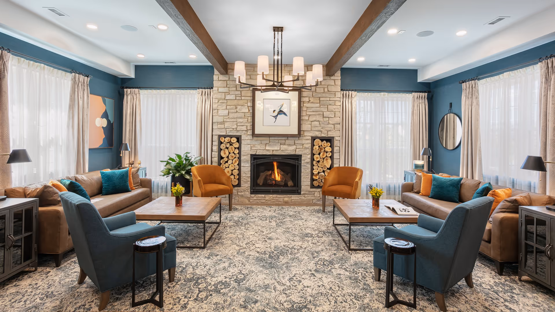 A cozy and well-lit living room with two brown leather sofas, two blue armchairs, and two orange armchairs arranged around two wooden coffee tables. The room features a stone fireplace with a fire burning, flanked by stacked firewood. The walls are painted blue with large windows covered by sheer white curtains and beige drapes. A modern chandelier hangs from the ceiling with wooden beams, and decorative elements include a framed bird artwork above the fireplace, a round mirror, and table lamps.