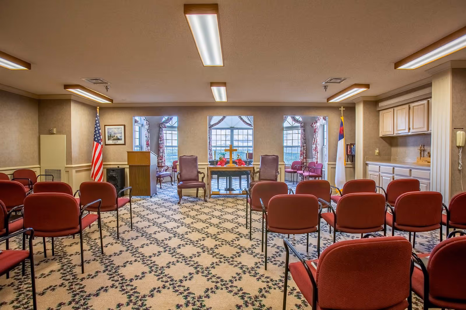 Interior view of a small chapel or meeting room with rows of red chairs facing a wooden podium and a table with a cross and flowers. The room has patterned carpet, three large windows with floral curtains, an American flag on the left, and a Christian flag on the right. There are two armchairs behind the table and cabinets on the right wall.