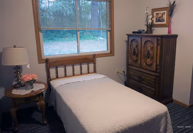 A small bedroom with a single bed covered in a white quilt, a wooden headboard, a wooden nightstand with a lamp and a vase of pink flowers, a window with blinds showing greenery outside, and a wooden dresser with decorative carvings and a framed picture on the wall above it.