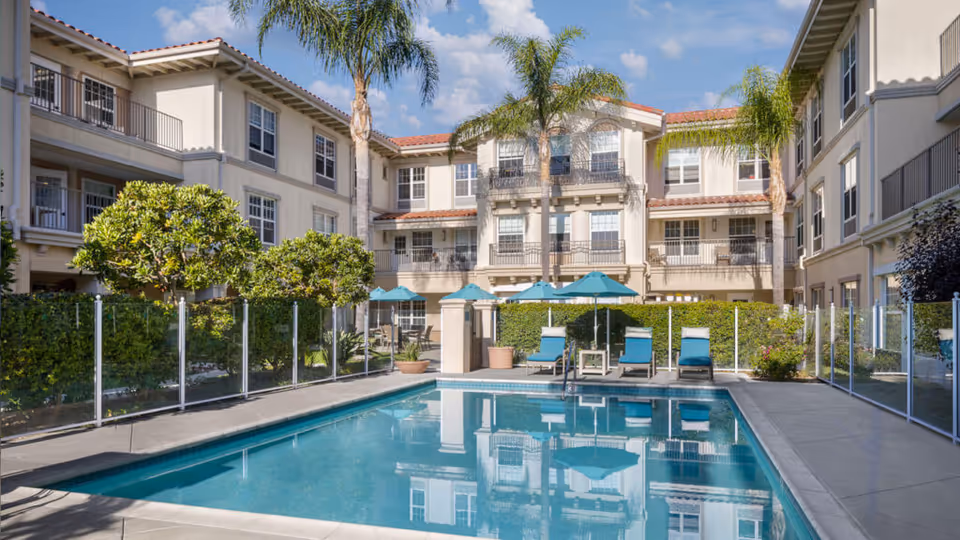 Outdoor swimming pool area at Belmont Village Senior Living Rancho Palos Verdes with lounge chairs, umbrellas, palm trees, and a three-story building in the background under a blue sky.