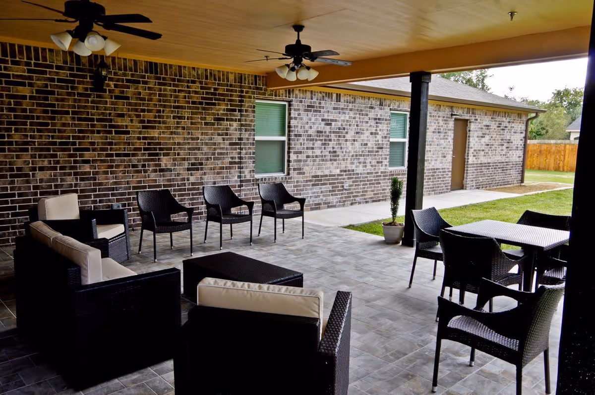 Covered outdoor patio area with black wicker chairs and tables, beige cushions, ceiling fans, and a brick wall in the background. The patio overlooks a grassy yard with a wooden fence.