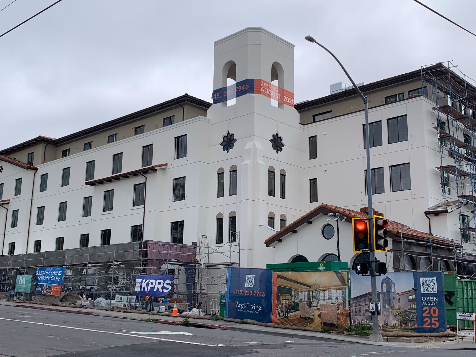Exterior view of a large multi-story building under construction with scaffolding and construction materials around. The building has a white facade with a tower featuring arched openings and decorative elements. Banners on the construction fence indicate it is an Aegis Living facility opening in August 2025. A traffic light and street signs are visible in the foreground.