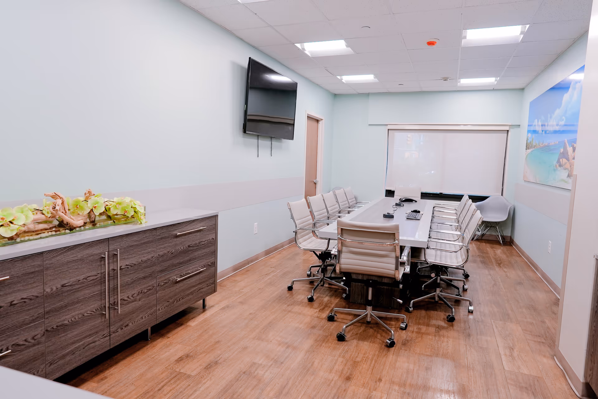 Conference room with a long table surrounded by swivel chairs, a wall-mounted TV, sideboard with plants, and a window with a blind.