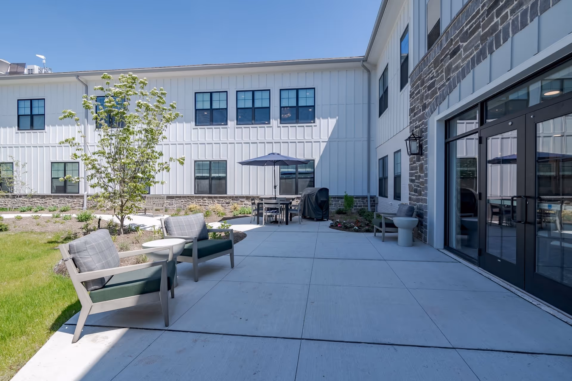 Outdoor patio area at Arbor Terrace Basking Ridge with two cushioned chairs and a small table on a concrete surface, a dining table with an umbrella, a grill, and a building with white siding and stone accents under a clear blue sky.