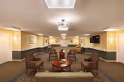 A well-lit assisted living facility common area with beige walls and carpeted floors. The room features a seating arrangement including four striped armchairs around a round wooden coffee table, a sofa in the foreground, framed artwork on the walls, and a flat-screen TV mounted on the right wall. Ceiling lights and wall sconces provide illumination.