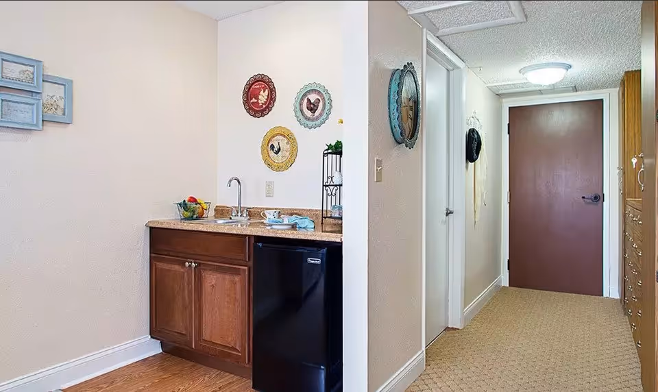 Interior view of a hallway and kitchenette area in a senior living facility. The kitchenette has a small sink, countertop, wooden cabinets, and a black mini-fridge. The hallway has a brown door at the end, beige walls, carpeted floor, and decorative wall hangings including colorful plates and framed pictures.
