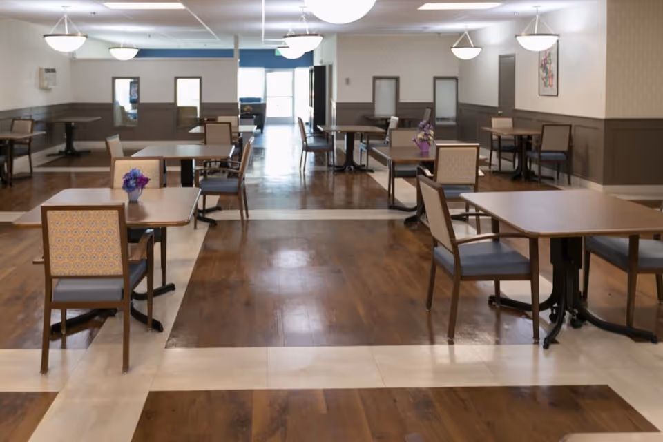 A spacious dining room with multiple square wooden tables and chairs arranged neatly. The floor has a combination of wood and light-colored tiles forming a pattern. The walls are painted in neutral tones with some framed artwork and windows. Ceiling lights provide bright illumination.