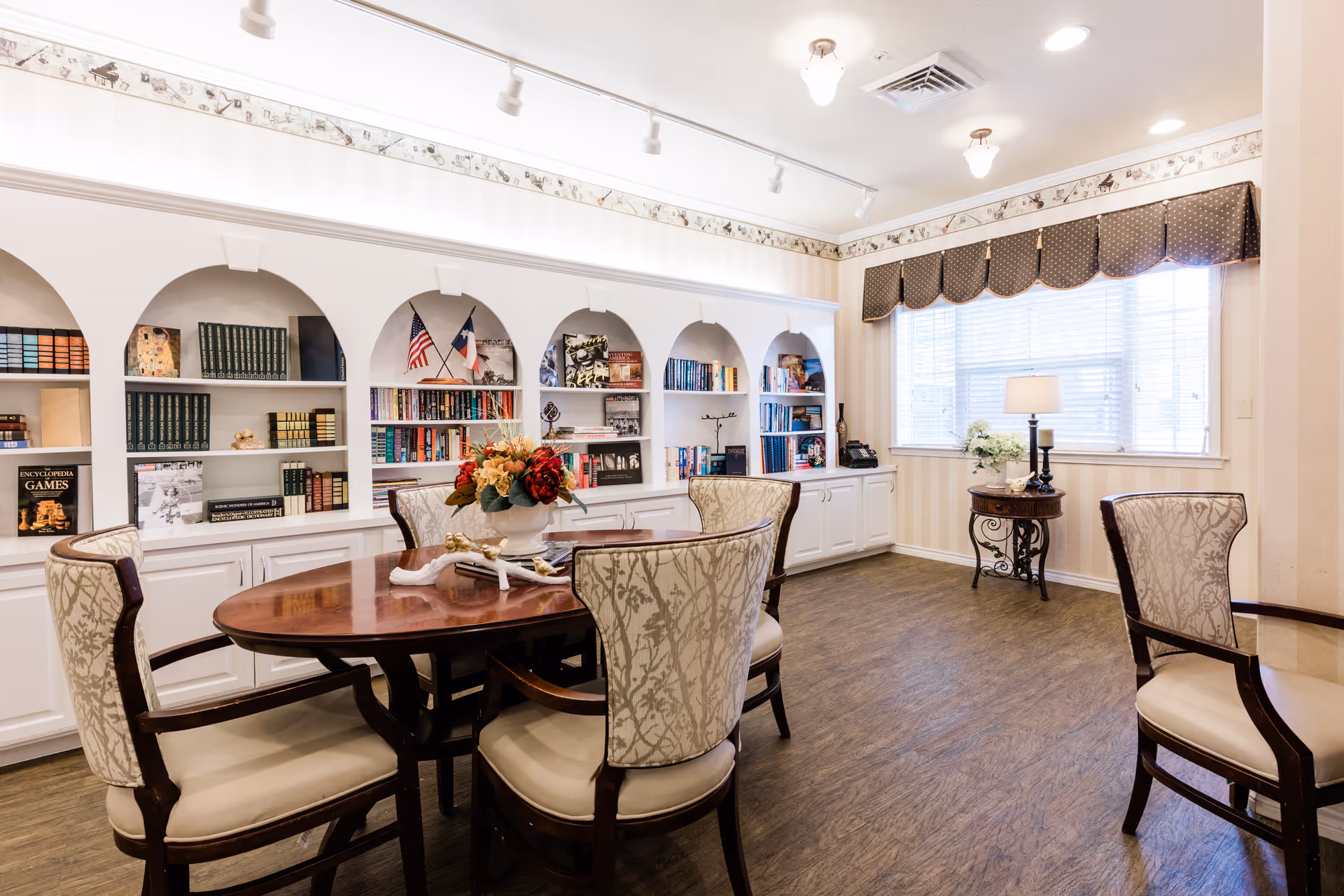 A bright and elegant room with a wooden round table surrounded by five upholstered chairs. The back wall features built-in white shelves with books, decorative items, and two small flags. A window with blinds and a valance lets in natural light, and a small round side table with a lamp and flowers is positioned near the window. The room has wood flooring and soft lighting from ceiling fixtures.