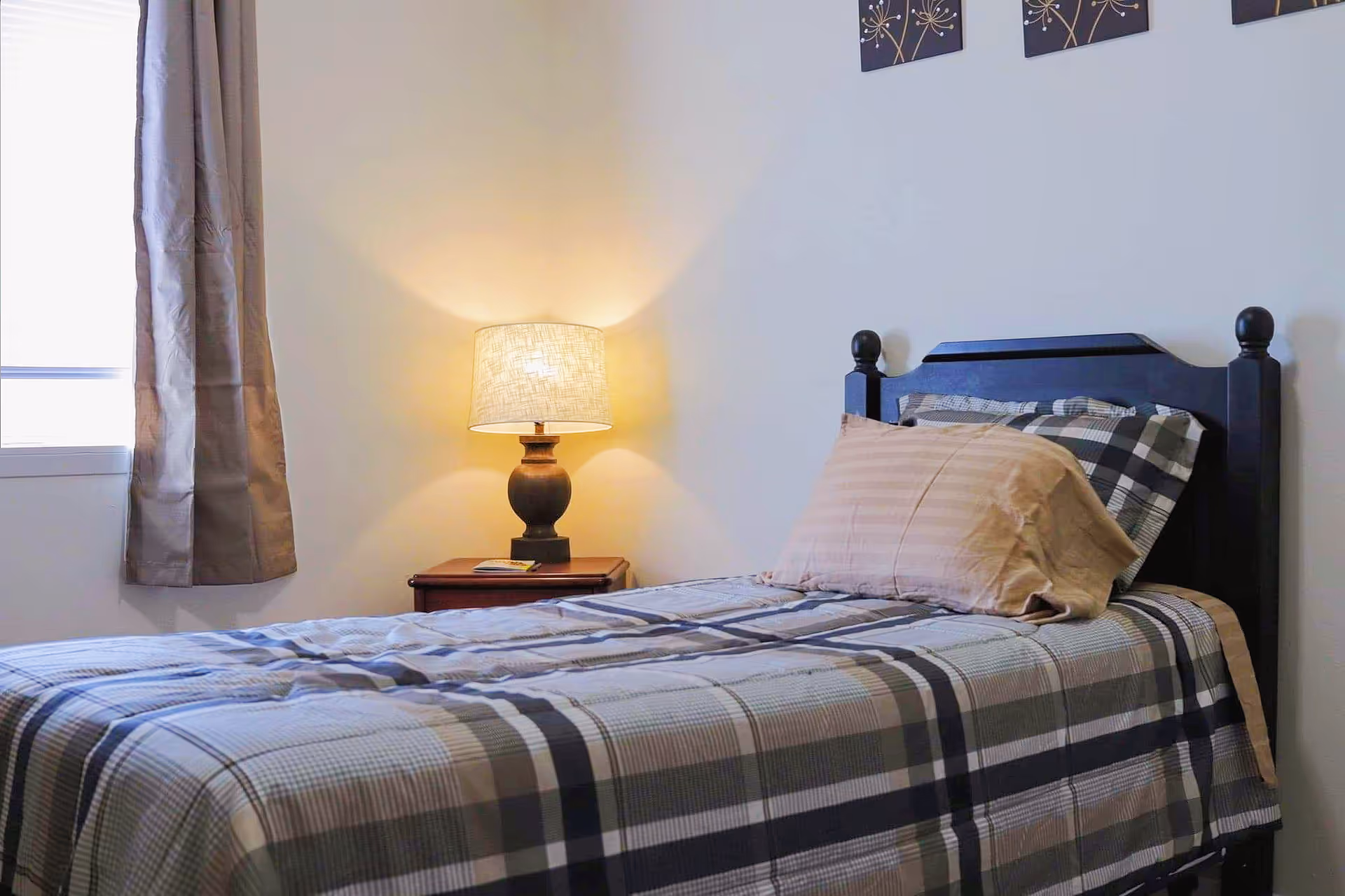 A neatly made single bed with plaid bedding and a beige pillow in a simple bedroom. Next to the bed is a wooden nightstand with a lit table lamp and a small book. A window with beige curtains is partially visible on the left, and two decorative wall hangings are above the bed.