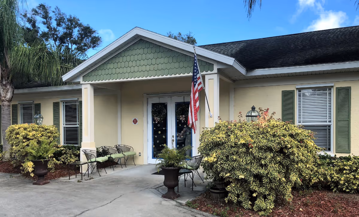 Front entrance of Spring Hills Assisted Living Facility showing a single-story building with a covered porch, two white doors decorated with wreaths, an American flag mounted on the right side, green shutters on windows, outdoor chairs with green cushions, and various bushes and plants around the entrance.