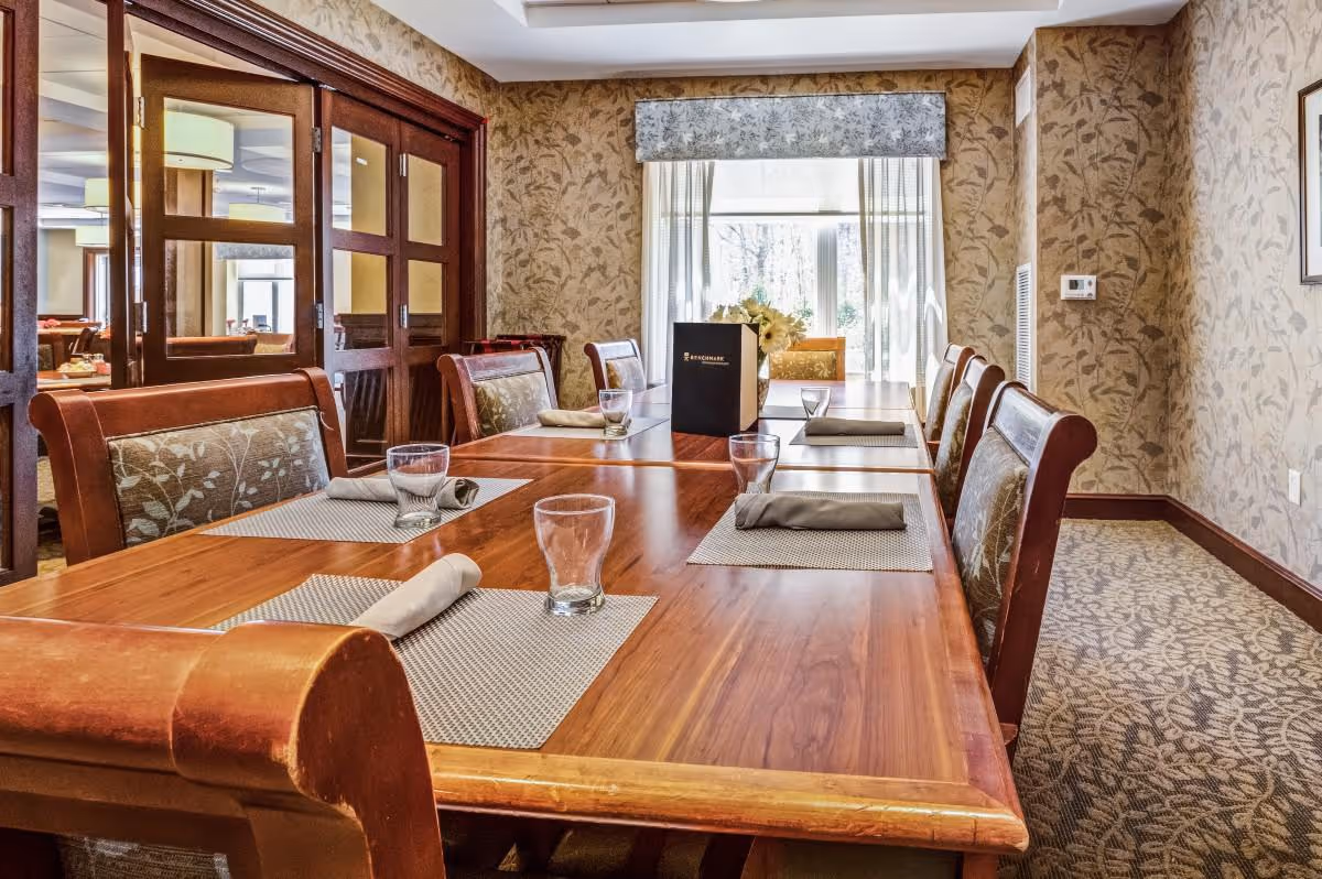 A dining room with a long wooden table set with placemats, rolled napkins, and empty glasses. The chairs have patterned upholstery. There is a window with sheer curtains and a floral valance at the far end of the room. The walls are decorated with floral wallpaper, and there is a wooden door with glass panels on the left side.