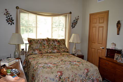 A cozy bedroom with a floral patterned bedspread and matching pillows on a bed positioned in front of a large window with blinds. Two bedside tables each hold a lamp with a beige shade. The room has light-colored walls, a wooden door, a dresser with framed photos, a doll, and decorative wall hangings.