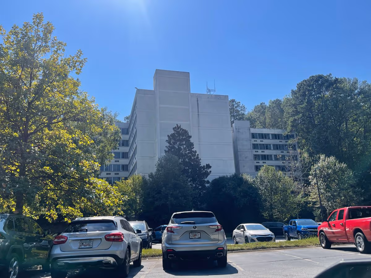 White multi-story residential building seen behind trees with a row of parked cars in the foreground under a clear blue sky.