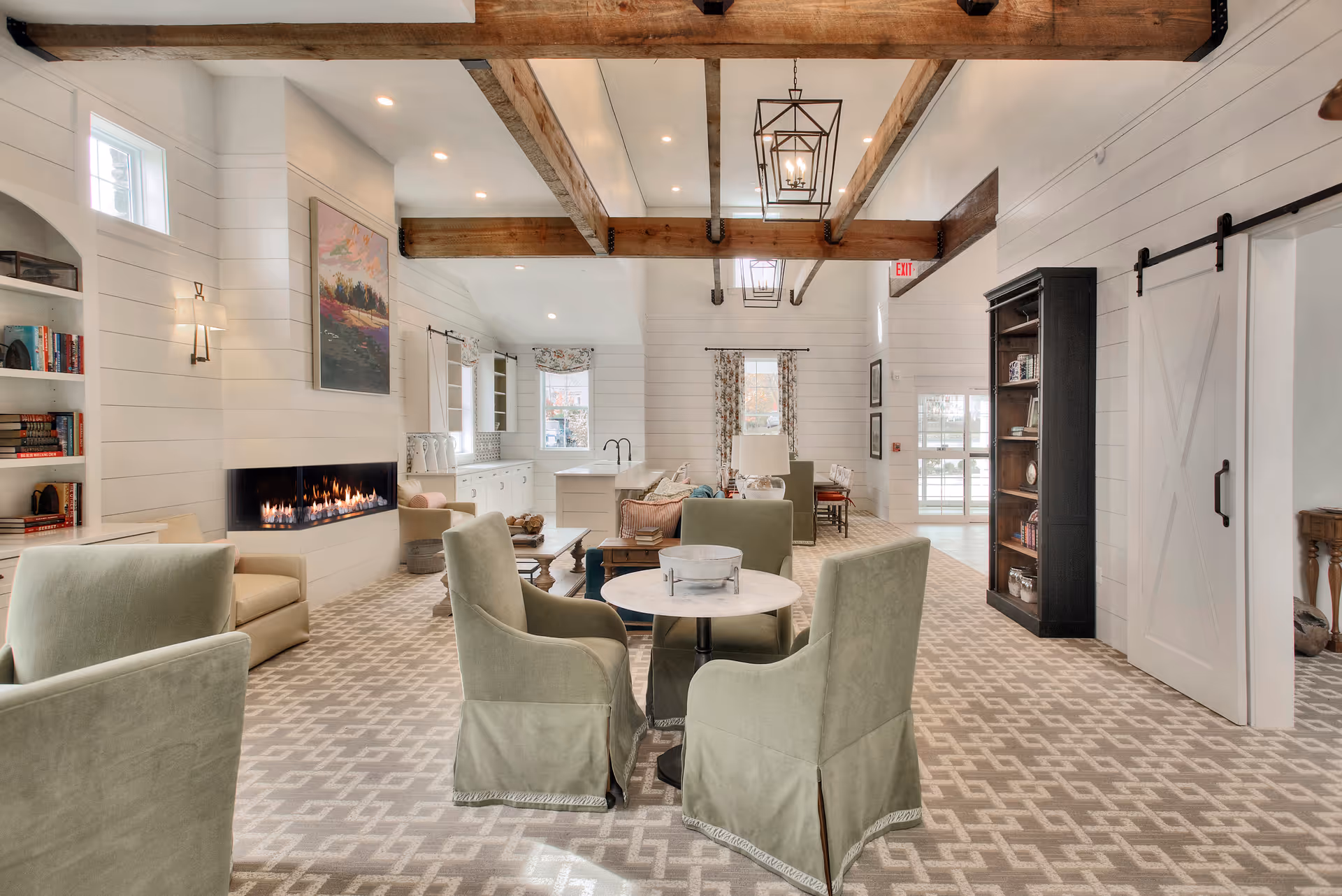 Bright open communal living area with upholstered chairs around small tables, a linear fireplace, built-in shelving and exposed wooden ceiling beams.