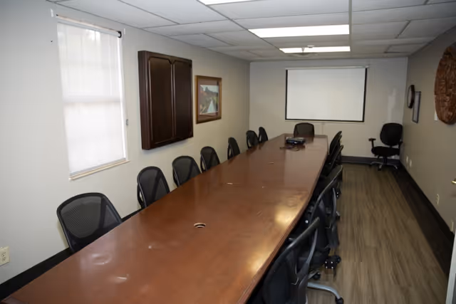 A long conference room with a large wooden table surrounded by black office chairs. The room has a window with a white blind, a wall-mounted cabinet, framed artwork, a projector on the table, and a white projection screen on the far wall. The floor is wood laminate, and the ceiling has fluorescent lighting.