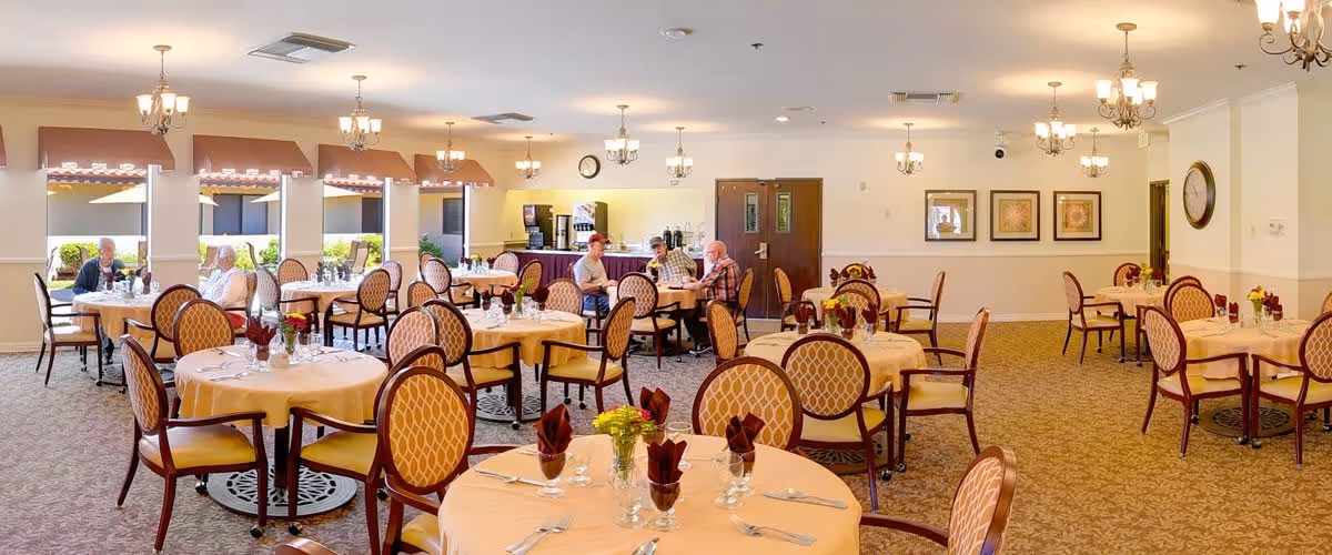 A spacious dining room with round tables covered in beige tablecloths, each set with silverware, glasses, and napkins. Several elderly people are seated at tables, engaged in conversation. The room has large windows with brown valances, chandeliers hanging from the ceiling, framed artwork on the walls, and a coffee station in the back.