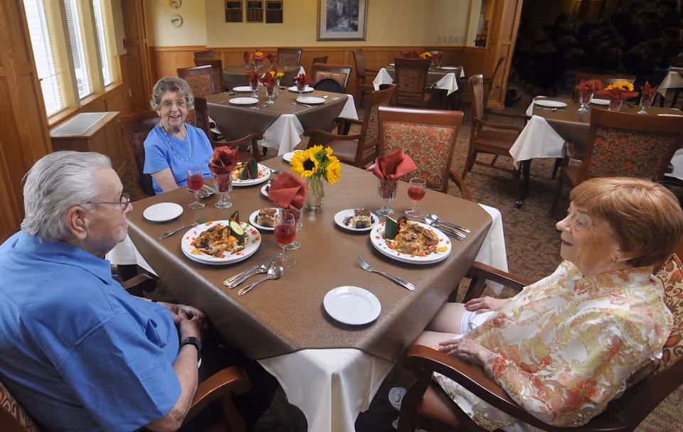 Three older adults seated at a dining table with plated meals, glasses and a sunflower centerpiece in a communal dining room.