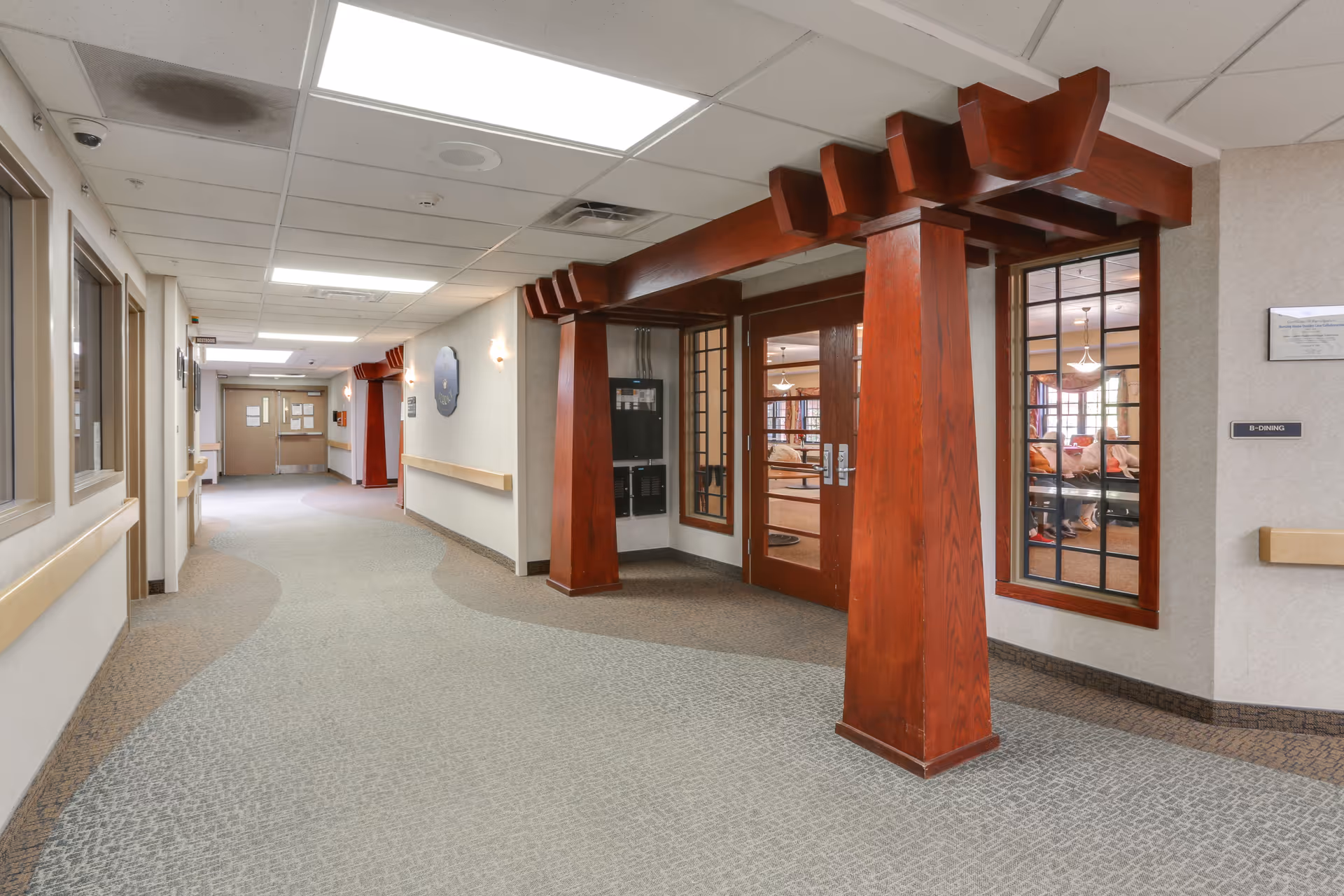 Interior hallway of a senior living facility with beige walls, carpeted floor, and wooden architectural accents around a set of double doors with glass panes. The hallway is well-lit with ceiling lights and has handrails along the walls. Through the glass doors, a common area with seating and people can be seen.
