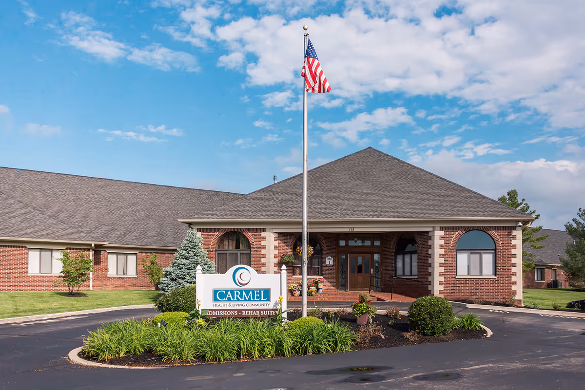 Front exterior of the Carmel Health & Living Community brick building with an American flag, landscaped entrance, and facility sign.