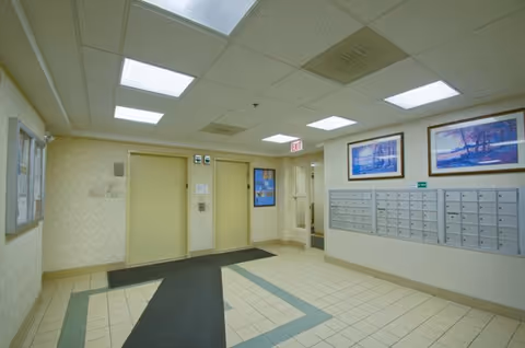 Interior view of a senior living facility hallway with two elevators, a bulletin board on the left wall, a row of mailboxes on the right wall, and two framed landscape paintings above the mailboxes. The floor is tiled with a patterned carpet runner leading to the elevators, and fluorescent ceiling lights illuminate the area.