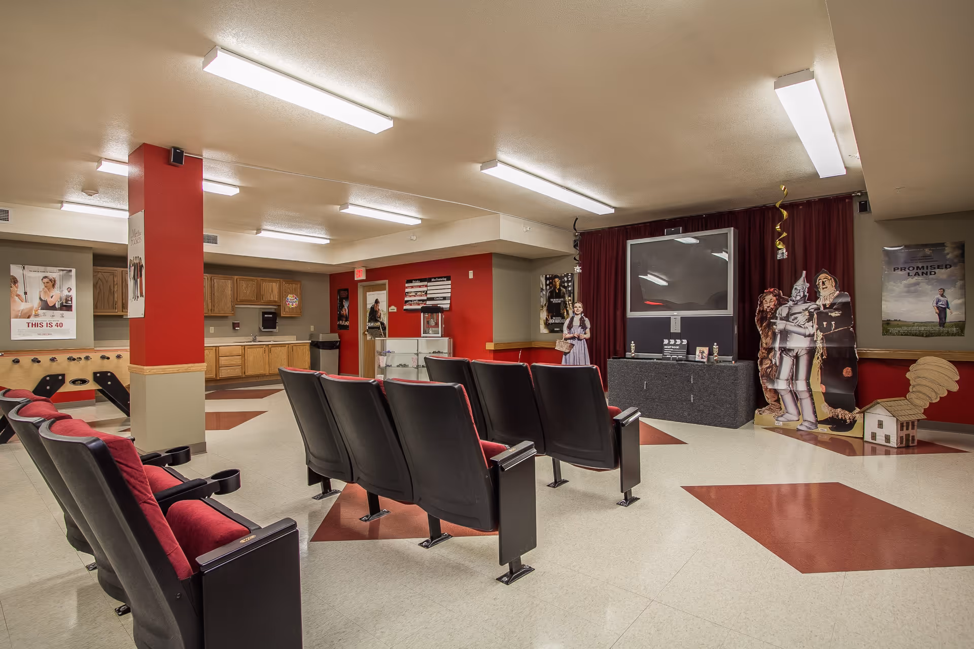 A small theater room with two rows of black and red cushioned seats facing a large flat-screen TV mounted on a dark gray cabinet. The room has a red and beige color scheme with red pillars and geometric patterns on the floor. There are movie posters on the walls and life-size cardboard cutouts of characters from The Wizard of Oz near the TV. The room also features a kitchenette area with wooden cabinets and a foosball table.