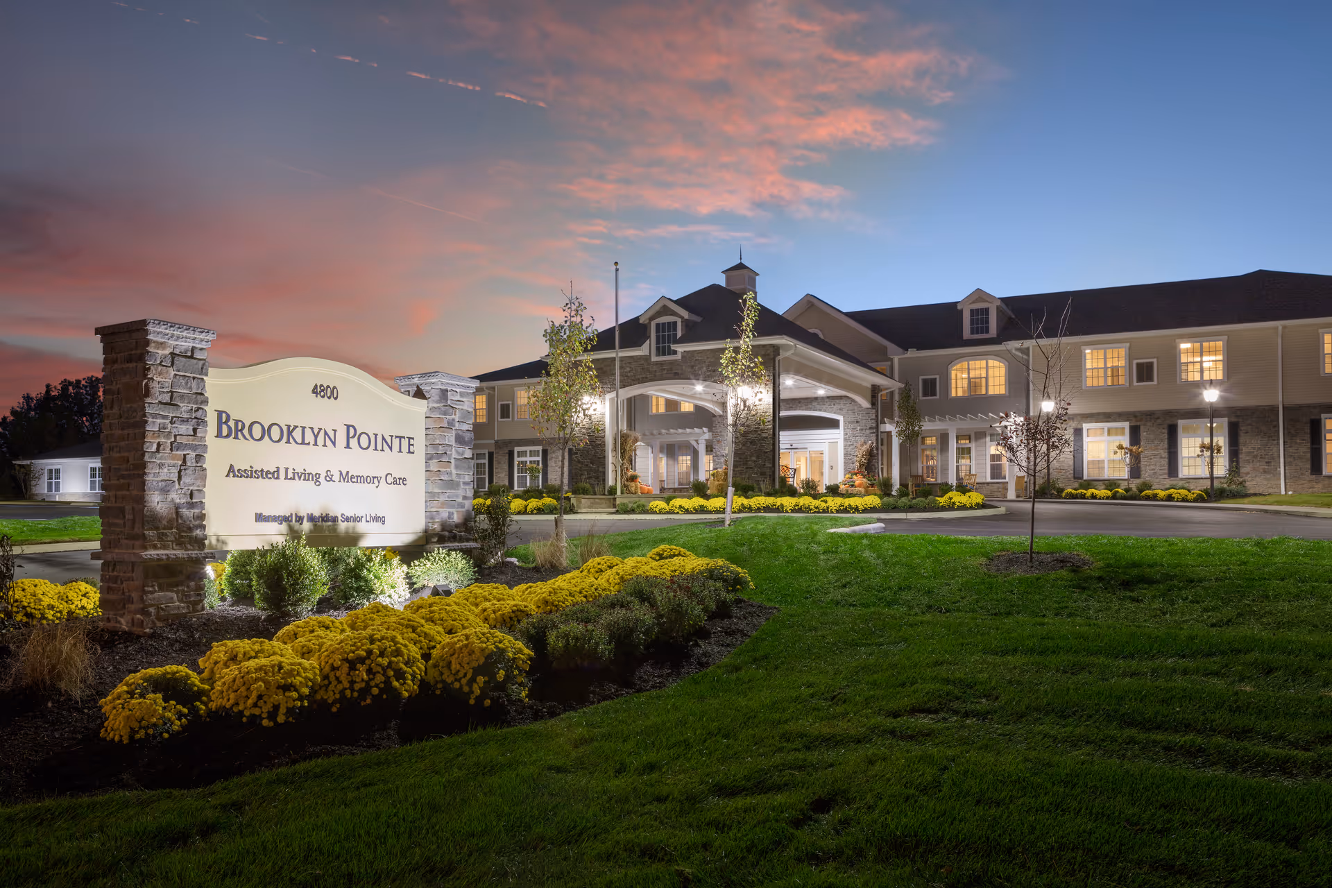 Front entrance of Brooklyn Pointe Assisted Living & Memory Care at dusk with an illuminated sign and landscaped grounds.