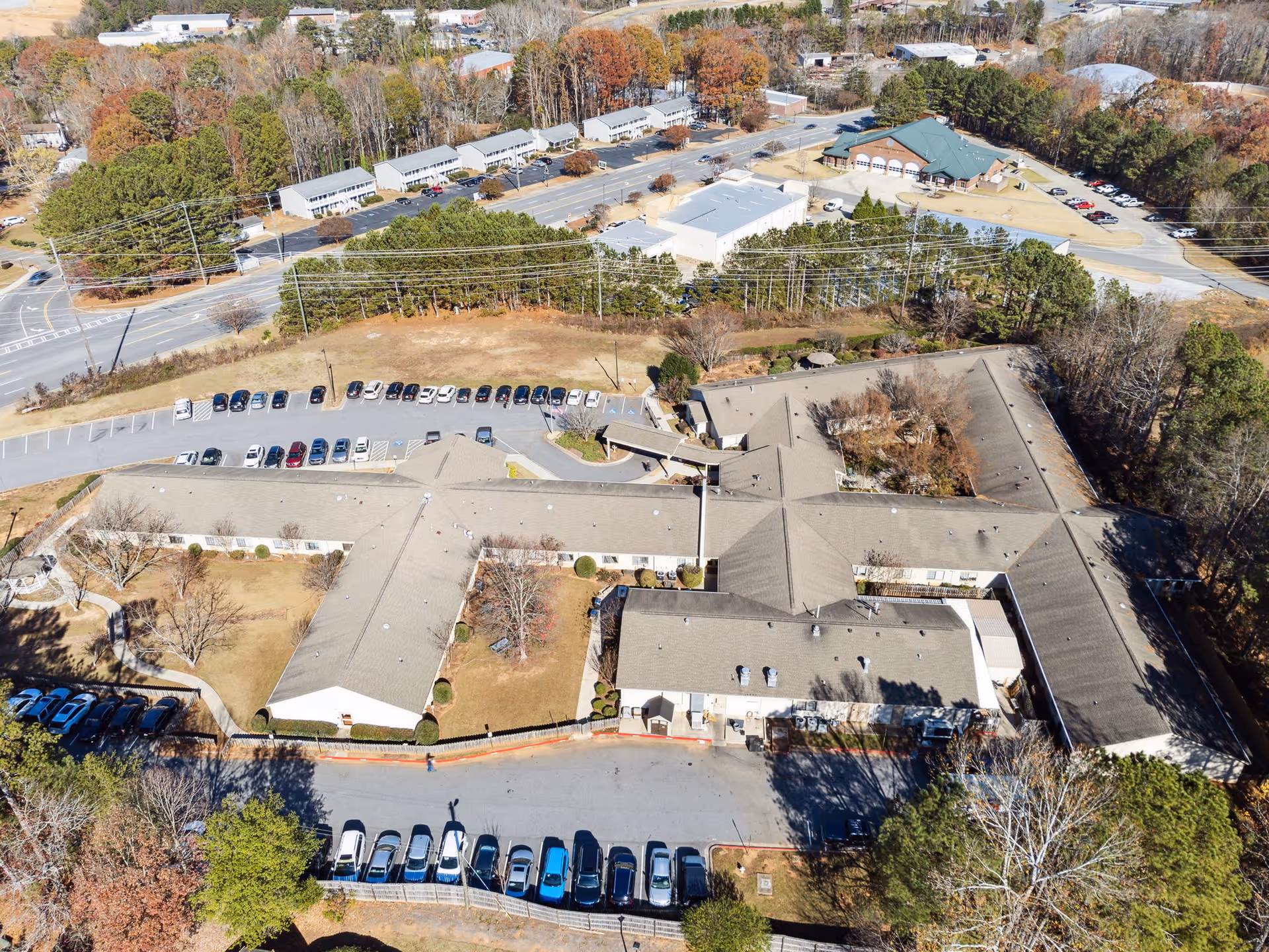 Aerial view of Cumming Health & Rehab facility showing a large building complex with multiple wings, surrounded by parking lots with numerous cars, trees, and nearby roads. The landscape includes some autumn-colored trees and open grassy areas.