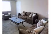 A living room with two brown sofas and one armchair arranged around a rectangular coffee table with a glass top. The room has beige walls, a carpeted floor, and a window with white blinds letting in natural light.