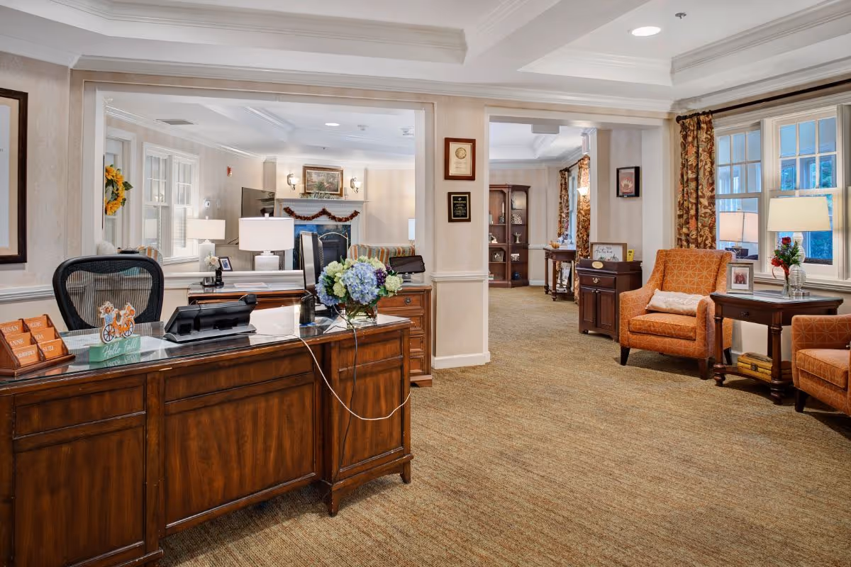 Reception area of a senior living facility with a wooden desk, office chair, and computer. The room features beige walls, carpeted floor, and large windows with floral curtains. There are two orange armchairs with a side table and lamp near the windows. In the background, a cozy living room area with a fireplace, TV, and framed artwork is visible.