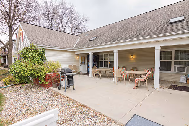 Outdoor patio area at Brookdale Mankato with a concrete floor, several tables and chairs with cushions, a barbecue grill, and a covered porch attached to a beige building. Leafless trees and some shrubbery are visible in the background.