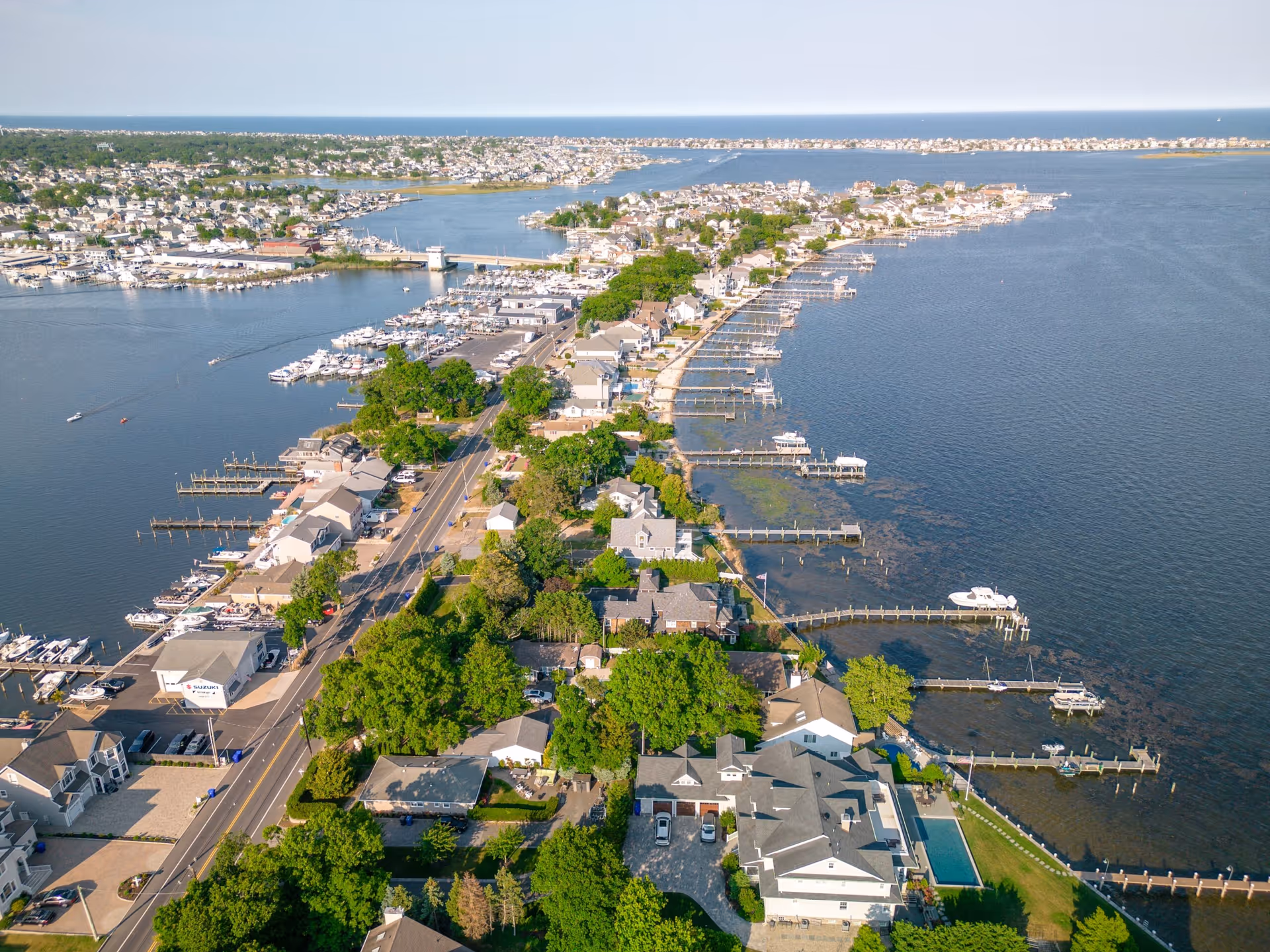 Aerial view of a coastal residential area with houses, docks, and boats along a narrow peninsula surrounded by water on both sides under a clear sky.