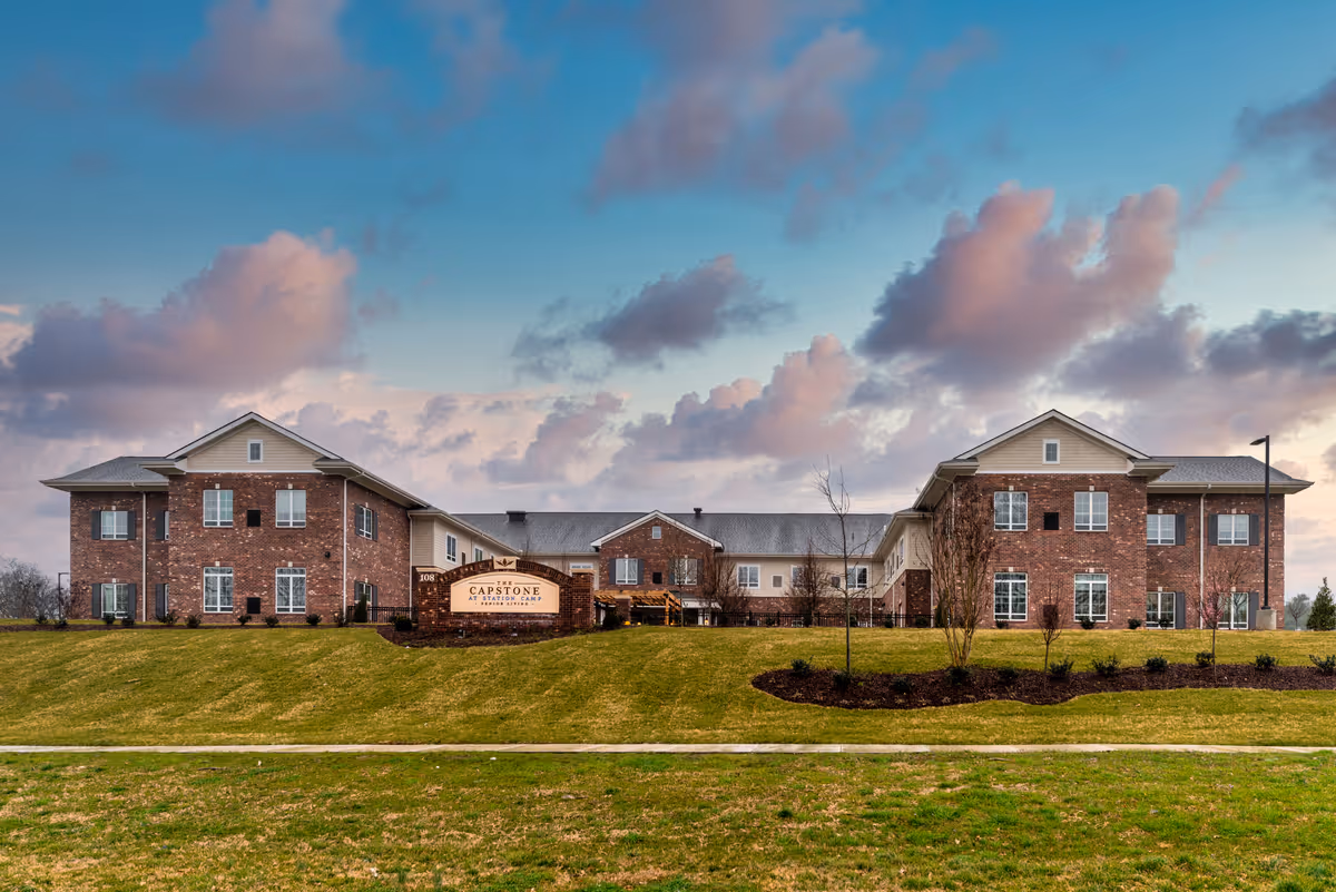 Exterior view of The Capstone at Station Camp, a large two-story brick building with multiple windows, surrounded by a well-maintained lawn and landscaping under a partly cloudy sky at sunset.