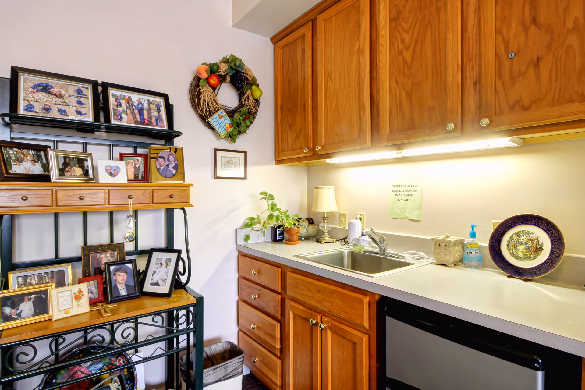 Interior view of a room with wooden cabinets and a countertop with a sink. On the countertop are a lamp, a potted plant, a bottle of hand sanitizer, a decorative plate, and a small ceramic container. Above the sink is a note reminding to drink 12 ounces in the morning and night. To the left, there is a metal and wood shelving unit displaying multiple framed family photos and a decorative wreath hanging on the wall.