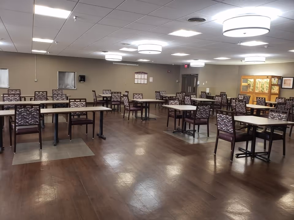 A spacious dining room with multiple tables and chairs arranged neatly on a wooden floor. The room is well-lit with ceiling lights and has beige walls. There is a display cabinet with plants or decorations on the right side and a bulletin board on the far wall.