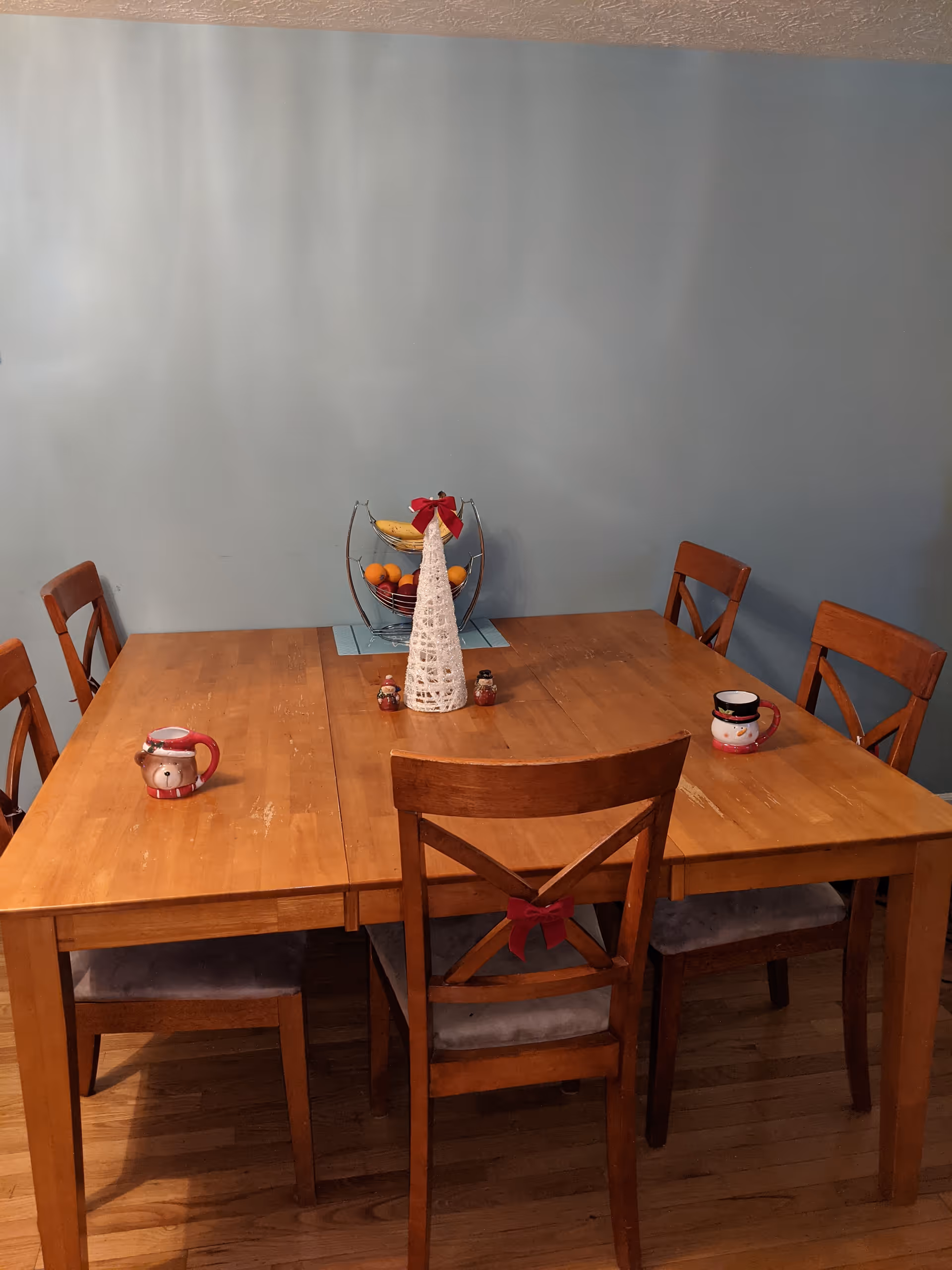 A wooden dining table with six matching wooden chairs in a room with light blue walls. On the table, there is a white decorative Christmas tree with a red bow, a two-tier fruit basket with bananas and oranges, two holiday-themed mugs, and two small decorative items. One chair has a red bow tied to its back.