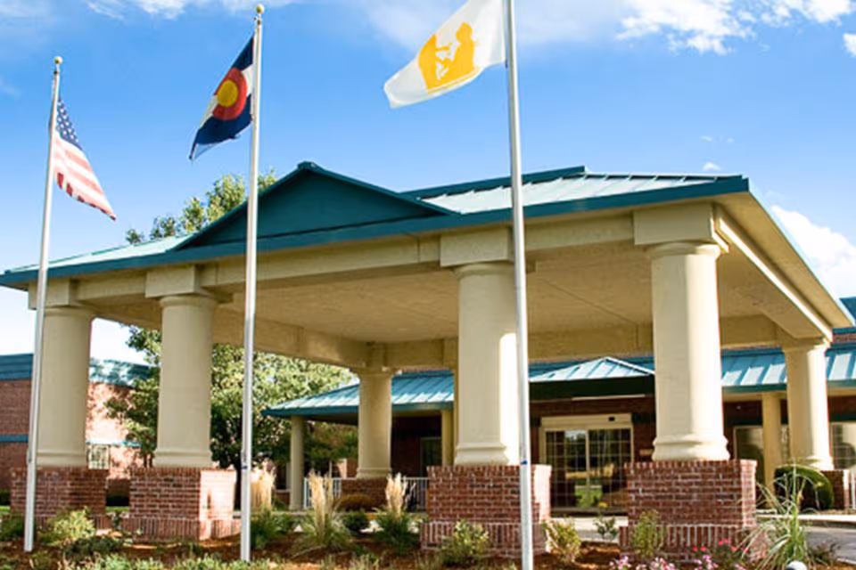 Entrance of a senior living facility with a covered driveway supported by large columns. Three flagpoles display the American flag, the Colorado state flag, and a white flag with a yellow logo. The building has a green roof and brick accents with landscaping in front.